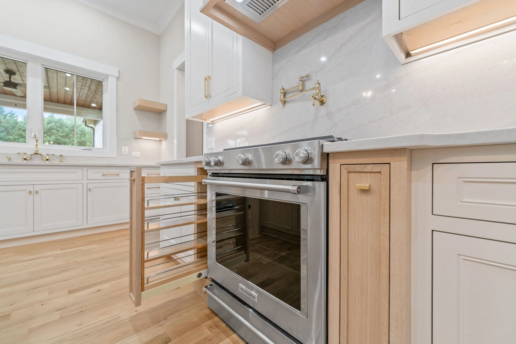 Luxury kitchen detail photo in Oak Ridge NC featuring KitchenAid stainless range, brass pot filler, marble backsplash, natural wood hood, and pull-out spice cabinet