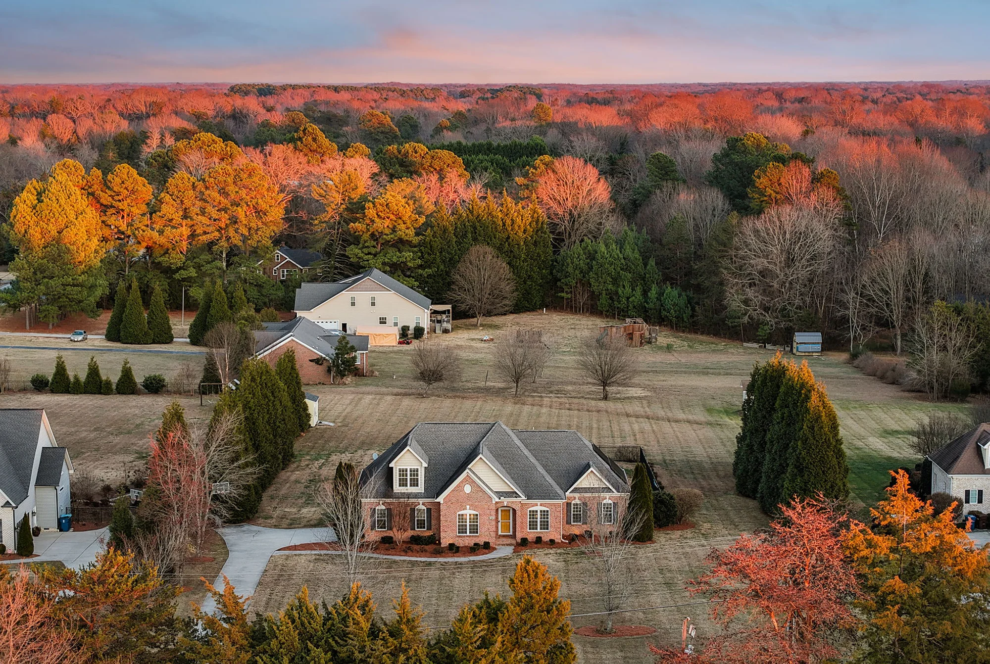 Aerial drone photo of brick home in Summerfield NC surrounded by wooded lot with fall foliage and sunset sky